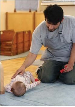 Un hombre sentado junto a un infante que está tirado en el suelo junto a él. El hombre está descansando su mano sobre el estómago del bebé y sosteniendo un juguete.