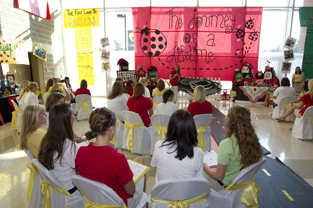 Cerca de uma dúzia de jovens mulheres são mostradas sentadas em cadeiras em uma irmandade de recrutamento no campus.