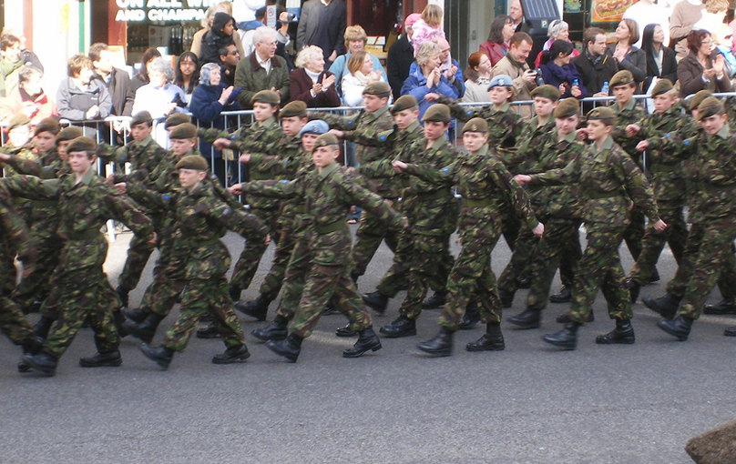 Un groupe de soldats est représenté en train de marcher sur la route sous les yeux des spectateurs.