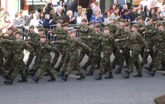 A group of soldiers is shown marching down the road while spectators look on.
