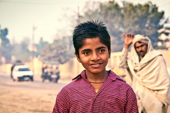 Young boy outside looking next to the camera while a man is waving in the back