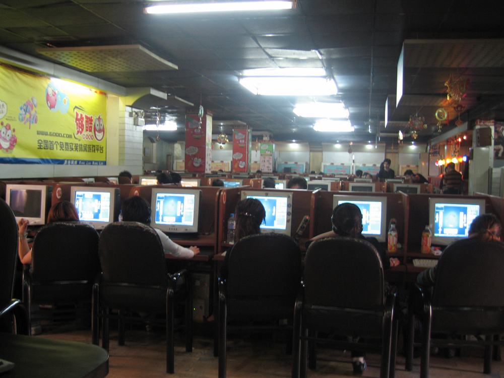 De nombreuses personnes assises sur des chaises regardent des écrans d'ordinateur dans un restaurant ou un café. Des affiches chinoises peuvent également être vues.