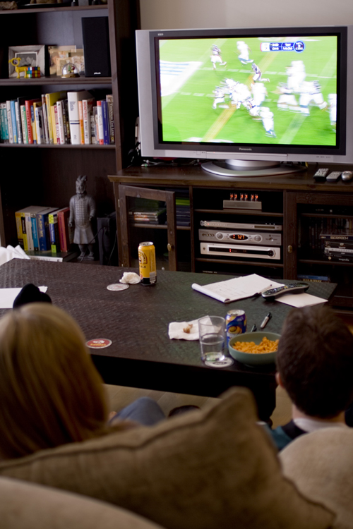 Un garçon et une fille sont montrés de derrière en train de regarder un match de football à la télévision. Une table basse se trouve entre eux et la télévision, et une étagère se trouve à côté du téléviseur.