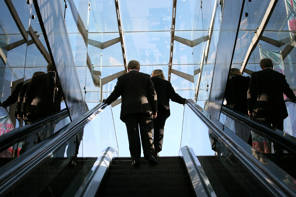 Un homme et une femme, tous deux vêtus de costumes d'affaires, sont représentés de dos en haut d'un escalator