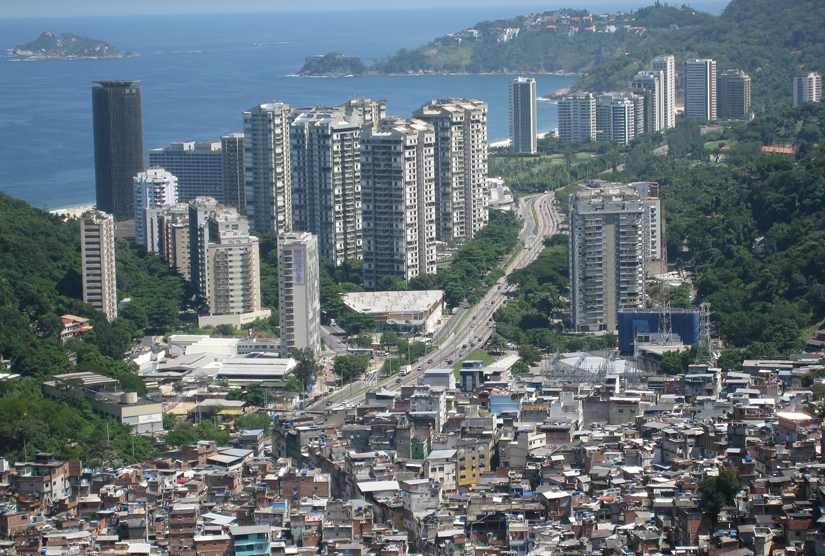 Esta foto é de uma cidade com grandes arranha-céus ao fundo e uma favela em primeiro plano.