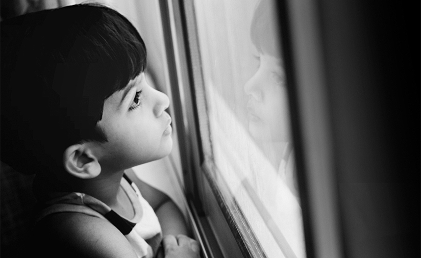 Cette photo représente un jeune garçon aux cheveux noirs regardant par la fenêtre.
