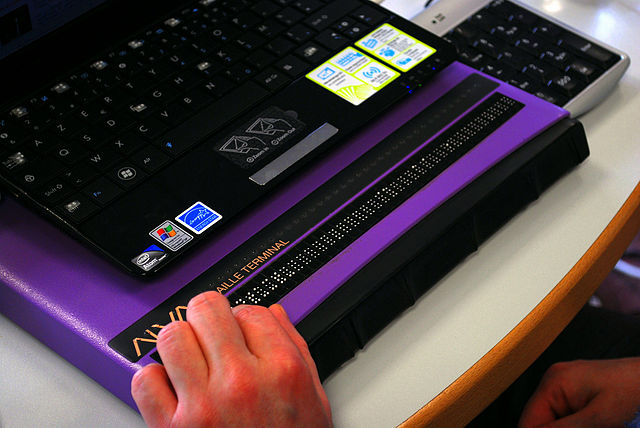 A refreshable Braille terminal device placed in front of a laptop, with a person's hand resting on the Braille.