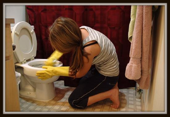 A woman is shown kneeling on bathroom floor scrubbing toilet.