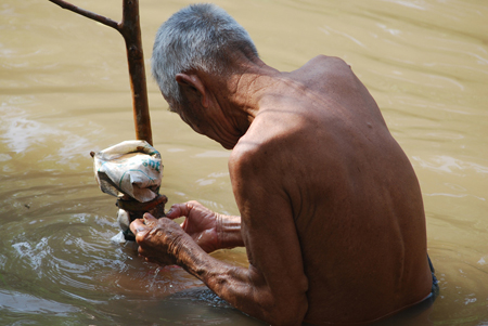 Un homme âgé torse nu est représenté en train de manipuler une grosse branche d'arbre alors qu'il se tient debout jusqu'à la taille dans une rivière.