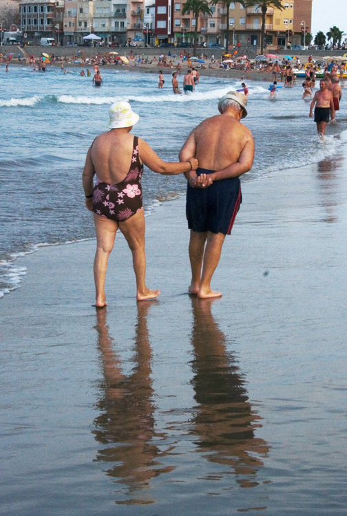 Un homme âgé portant un maillot de bain bleu et une femme âgée portant un maillot de bain fleuri et un chapeau sont représentés marchant au bord de l'eau sur une plage.
