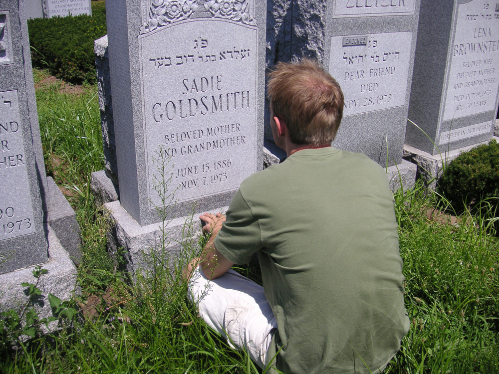 Un jeune homme vêtu d'un t-shirt vert et d'un short blanc est représenté assis dans l'herbe devant une pierre tombale.