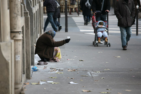 في الشكل (ب)، يظهر شخص بلا مأوى، يرتدي ملابس متهالكة، جالسًا على رصيف المدينة، حاملاً كوبًا بلاستيكيًا، يتوسل للتغيير من المارة.