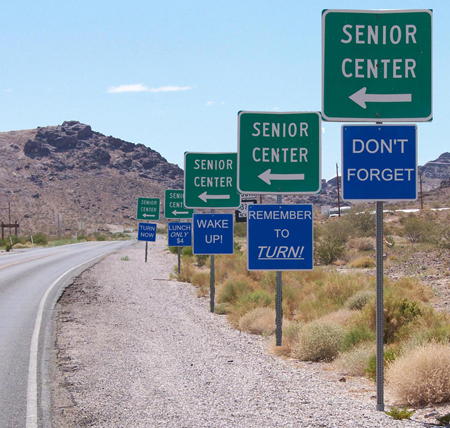 Cinq séries de panneaux de signalisation, la partie supérieure verte et la partie inférieure rouge dans chaque série, sont affichées sur le côté droit d'une route dans un environnement désertique. Les panneaux verts indiquent tous « Senior Center » et comportent une flèche pointant vers la gauche. Les panneaux bleus, de l'avant vers l'arrière, indiquaient « N'oubliez pas », « N'oubliez pas de [u] tourner ! [/u] », « Réveillez-vous ! » , « Déjeuner seulement 4$ » et « Tournez maintenant ».