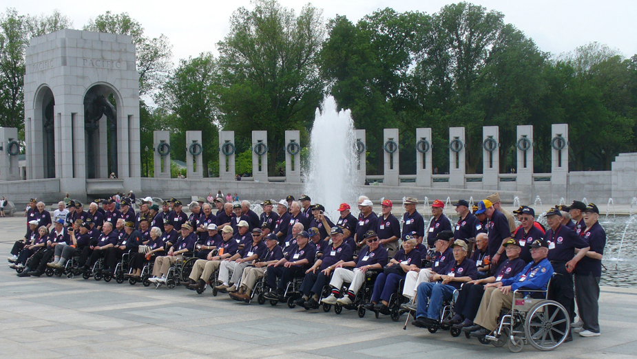 Um grupo de homens idosos, muitos em cadeiras de rodas, todos vestidos com camisas azuis e bonés de beisebol, são mostrados em pé e sentados em um ambiente memorial, com uma fonte e pilares atrás deles.