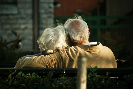 An elderly man and woman are shown from behind sitting on a bench. The man is shown wrapping his arm around the woman’s shoulders.