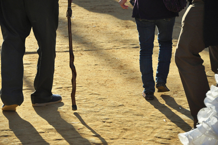 As pernas de três homens, um usando uma bengala, são mostradas por trás caminhando sobre uma superfície de terra.