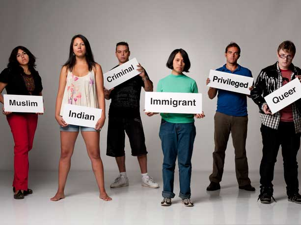 A group of people stand holding signs labeling them as others perceive them. The signs include, "Muslim", "Indian", "Criminal", "Immigrant", "Privileged", and "Queer".