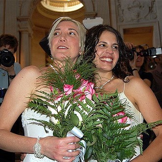 A bride and bride dressed in traditional white wedding gowns hold bouquets of flowers and smile for photos after a wedding ceremony.