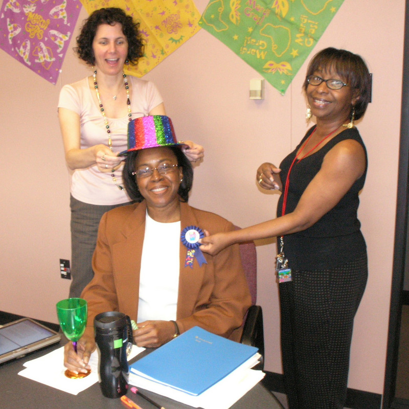 A pair of coworkers put a colorful party hat and ribbon on a colleague as part of an office birthday celebration.