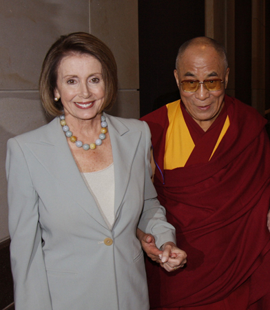 A foto mostra, à esquerda, a líder da minoria na Câmara, Nancy Pelosi, vestida com um terno cinza, de mãos dadas com, à direita, Dalai Lama Tenzin Gyatso, vestida com vestes marrons e amarelas.