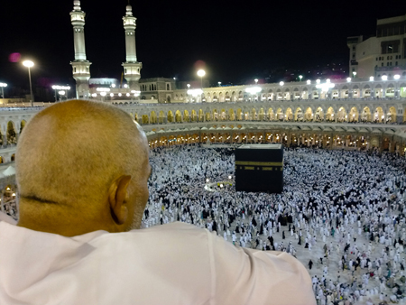 Um homem vestido de branco é mostrado por trás olhando para a Caaba, o local mais sagrado do Islã. Centenas de outras pessoas, vestidas de preto ou branco, podem ser vistas circulando uma grande estrutura em forma de cubo preto no chão de uma estrutura semelhante a um estádio.