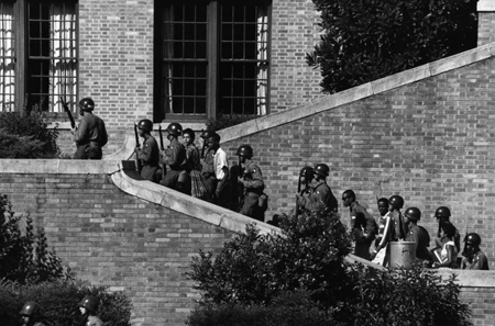 Des gardes nationaux armés escortent des étudiants noirs dans les escaliers extérieurs d'un lycée en brique.