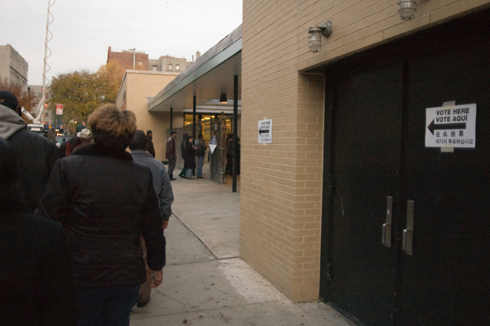 Des personnes font la queue devant un bâtiment. Des panneaux sur le bâtiment indiquent « Votez ici » en plusieurs langues.