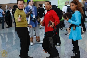 Three people in costume pose in a convention hall, holding prop weapons. The background features blurred attendees and displays.