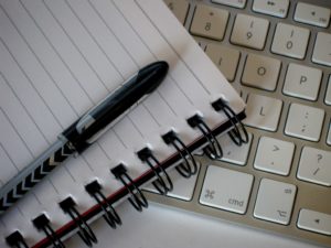 Image of a black pen on top of an open spiral notebook on top of a white keyboard.