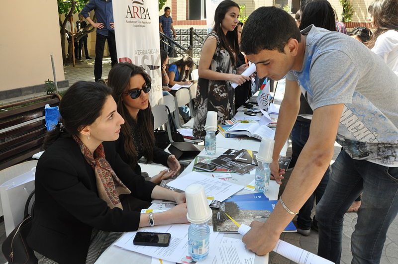 Photo of an outdoor information fair. In the foreground, a young man leans over a table where two women sit, papers, catalogs, and water bottles spread out in front of them.