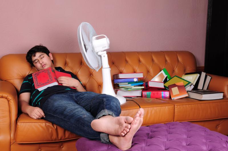 Photo of a male student asleep on a couch amid a pile of books. A fan is aimed at his face, and a book lies open on his chest.