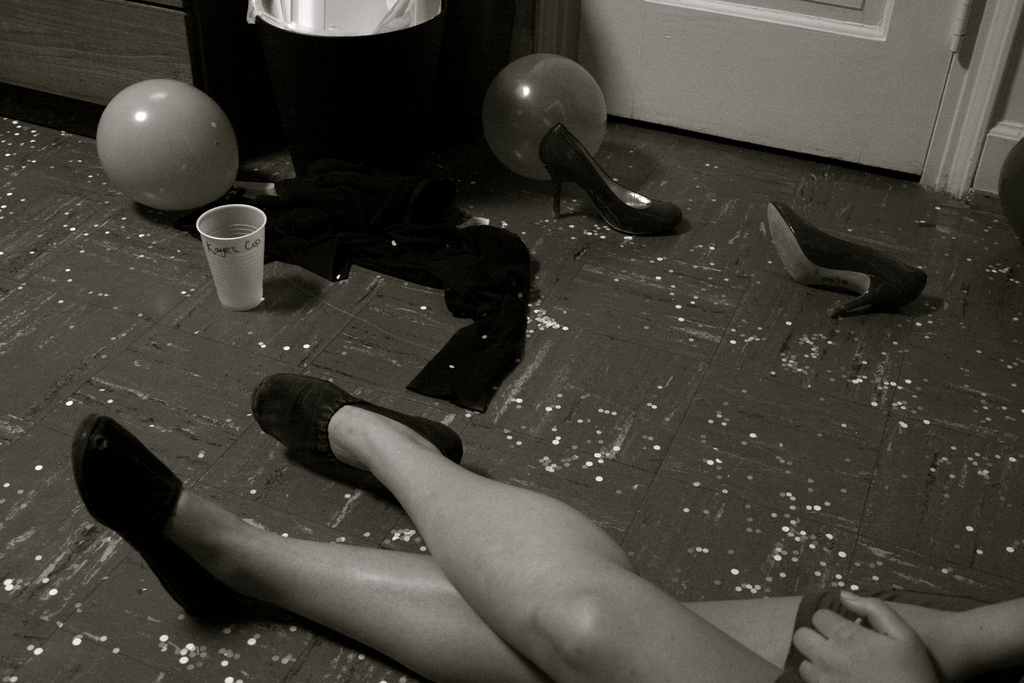 Photo of a woman's legs. Though mostly out of the frame, she appears to be sitting on the floor at a party. Next to her, on the floor, is a pair of high-heeled shoes, a party balloon, a plastic beer cup.