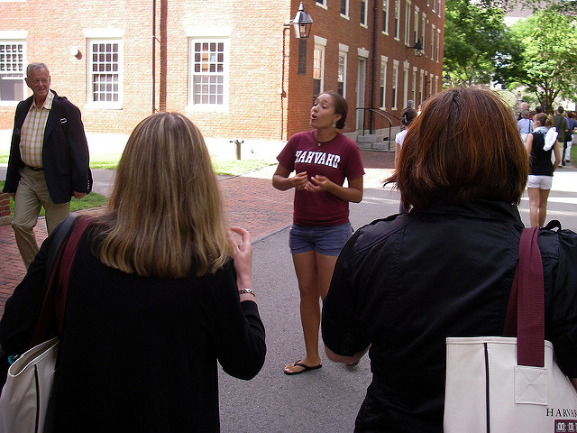 Campus tour guide wearing a shirt reading 'HAHVAHD" gesturing in front of a group of people, in front of a campus building