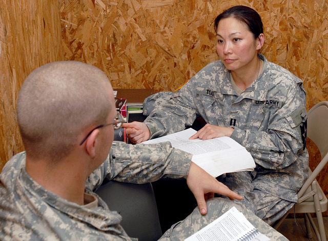 A female captain in the army reads a medical textbook at her desk while conversing with another soldier.