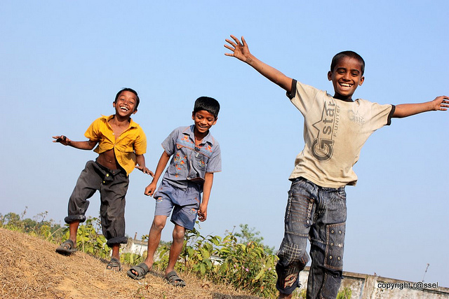 Three young boys playing outside and smiling widely. One boy looks directly at the camera, smiling with his arms outstretched.