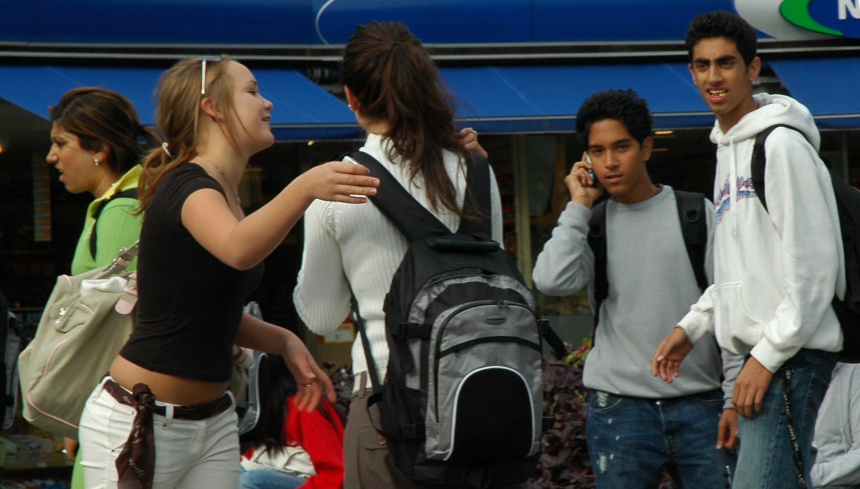 Youth from diverse backgrounds interacting and talking. Some are wearing backpacks, and one is talking on a cell phone.