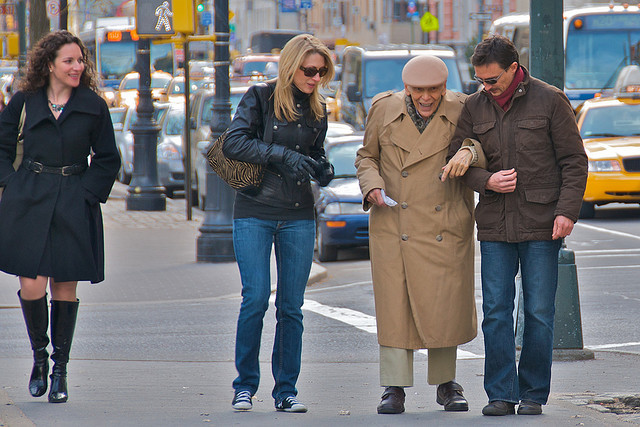 A man walks arm in arm with an elderly man, while another woman approaches as if to offer help as well. Yet another woman walks by smiling at the altruistic behavior.
