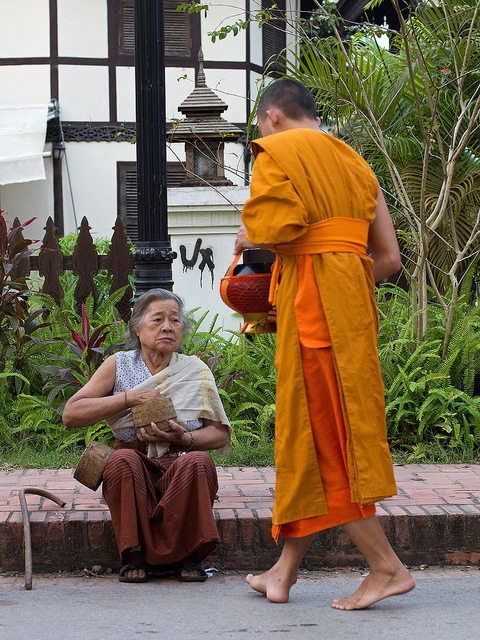 Photo of an older woman sitting on the sidewalk, reaching in her purse to give money to a monk.