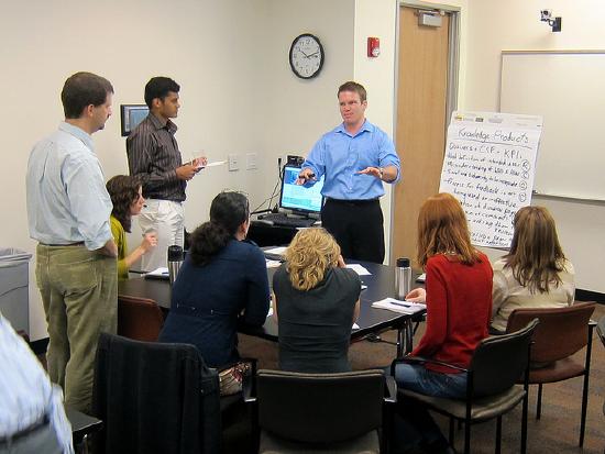 Team meeting. A man stands at the front of the room and his co-workers sit and stand around in a semi-circle.