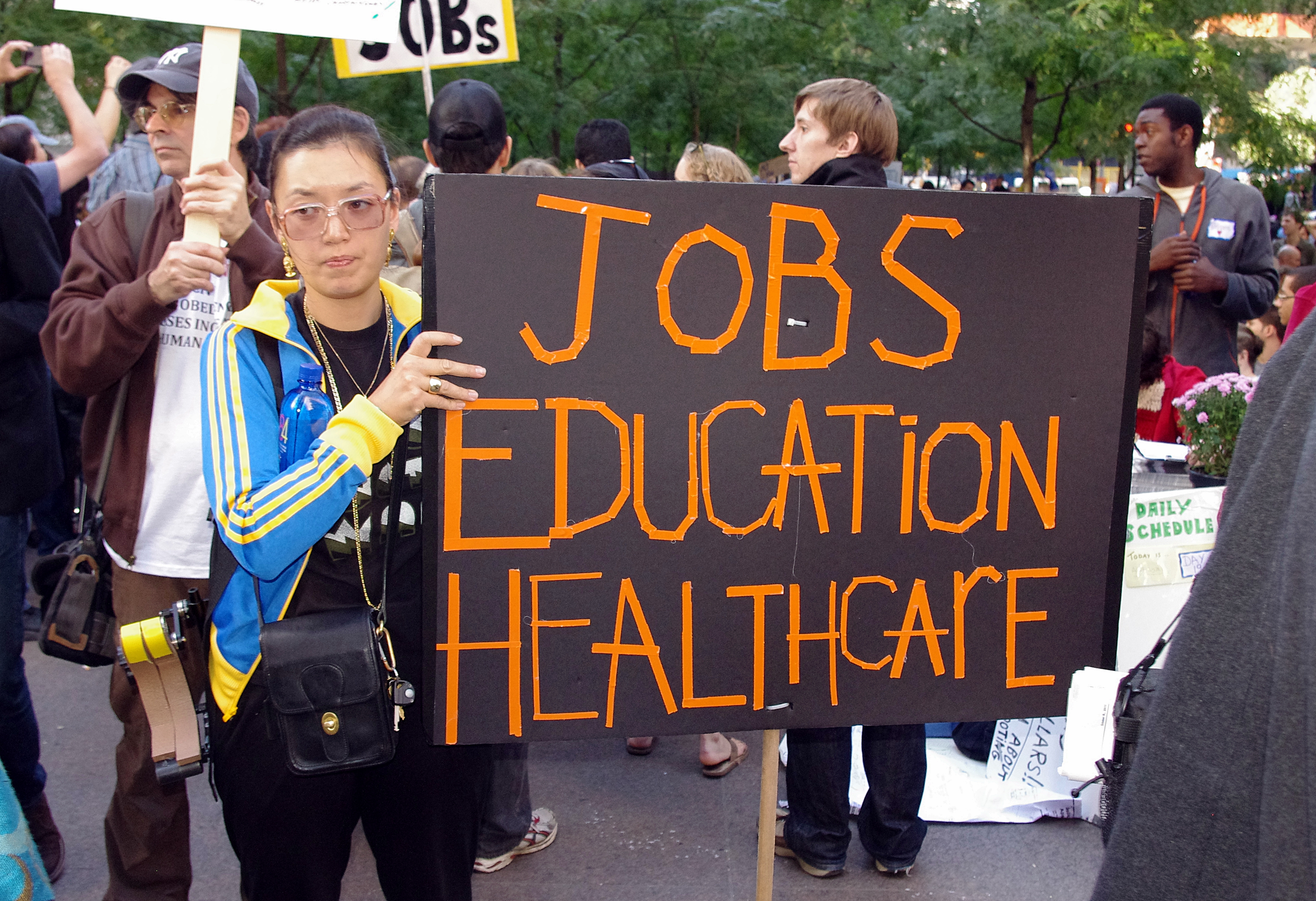 A woman stands in the foreground holding a protest sign that reads "Jobs, Education, Healthcare". There are other protestors in the background.