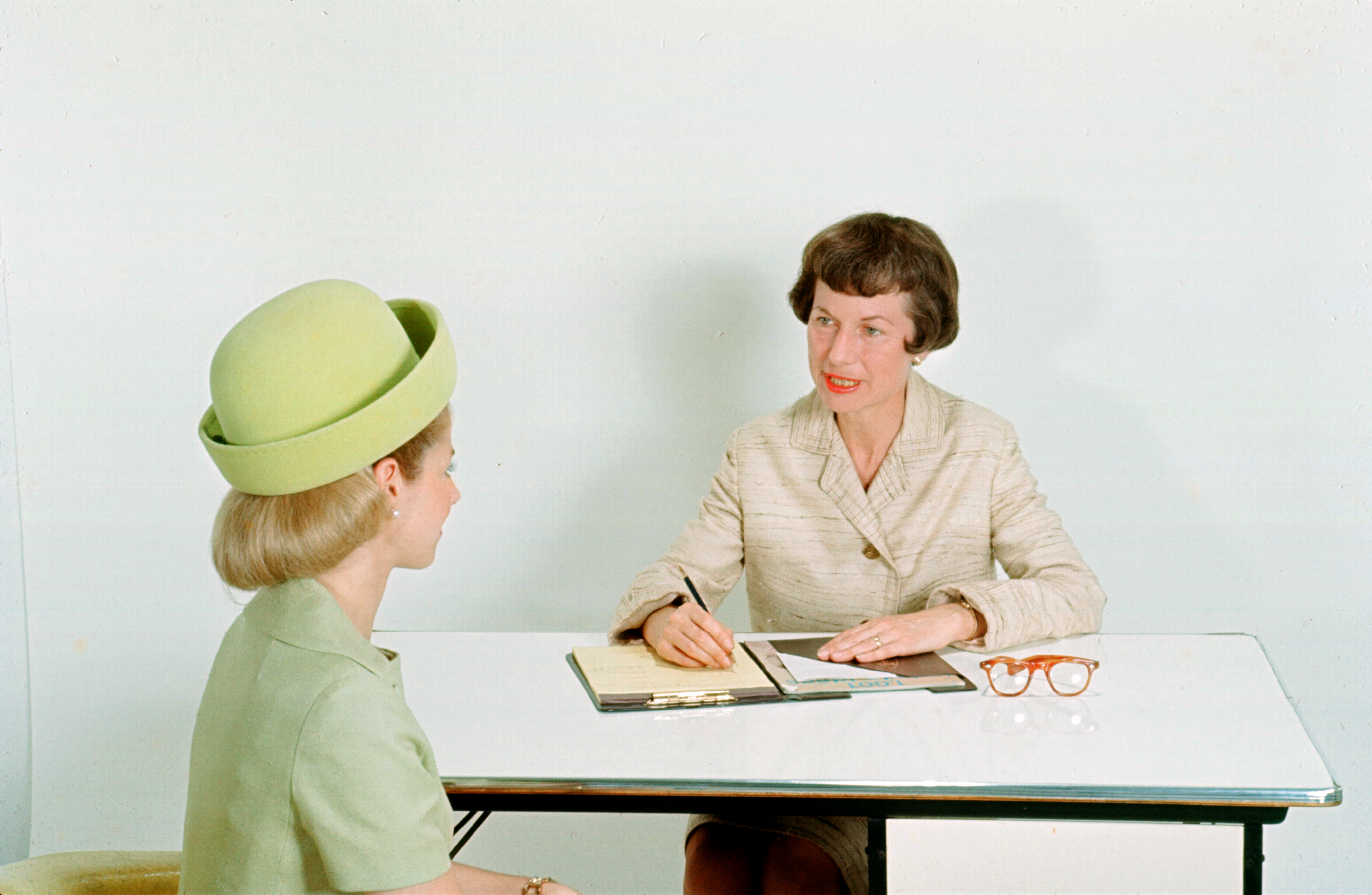 A woman sits behind a desk in professional attire, talking to a second woman and making notes in her notebook. The second woman is in the foreground and facing away from the camera and looking at the woman behind the desk.