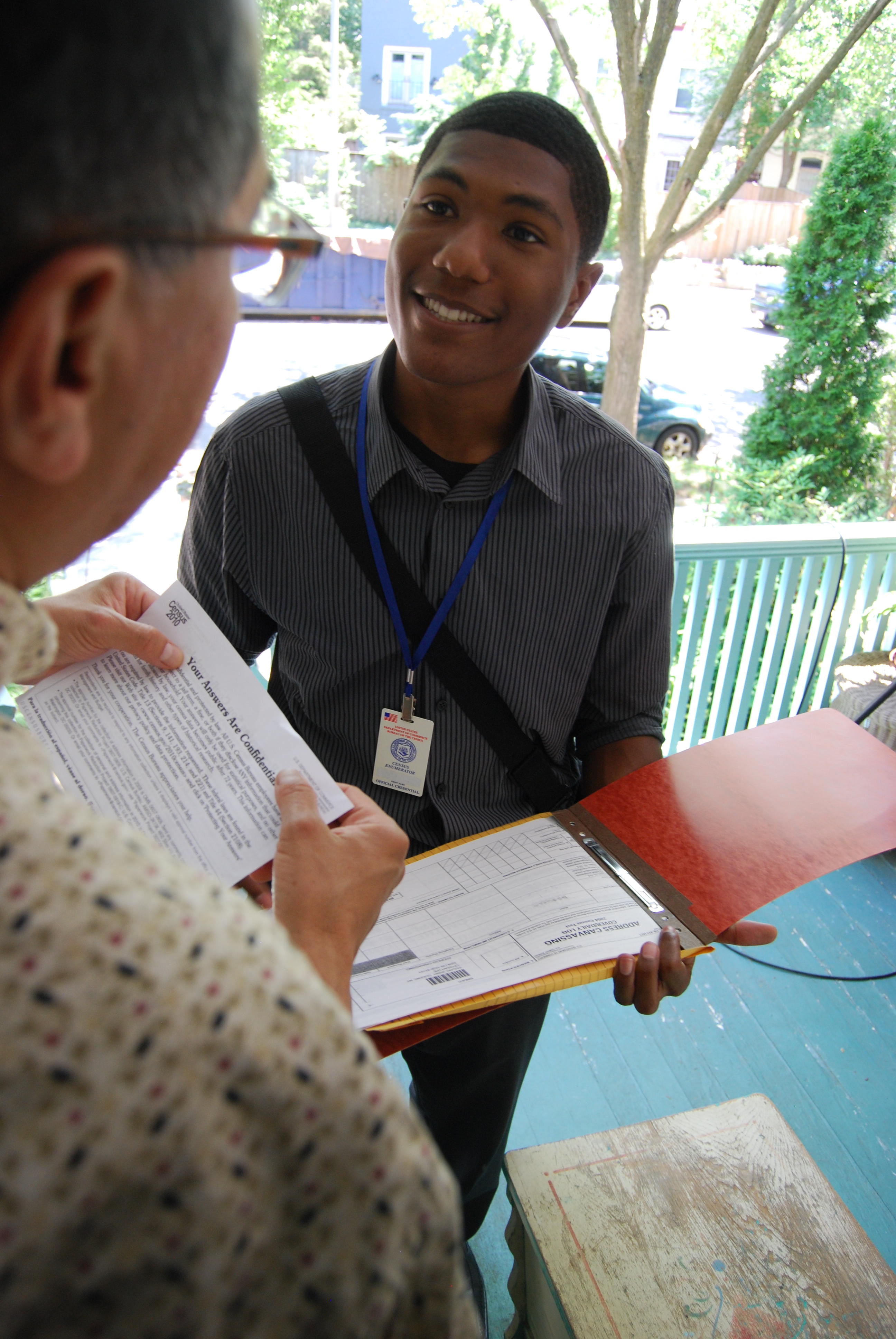 A man with a census enumerator badge is standing on another man's porch holding an open folder with papers in it. A second man, wearing glasses, is in the foreground looking at the census worker standing on his porch.
