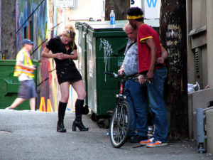 A woman injecting her arm in an alleyway while two other men stand nearby.
