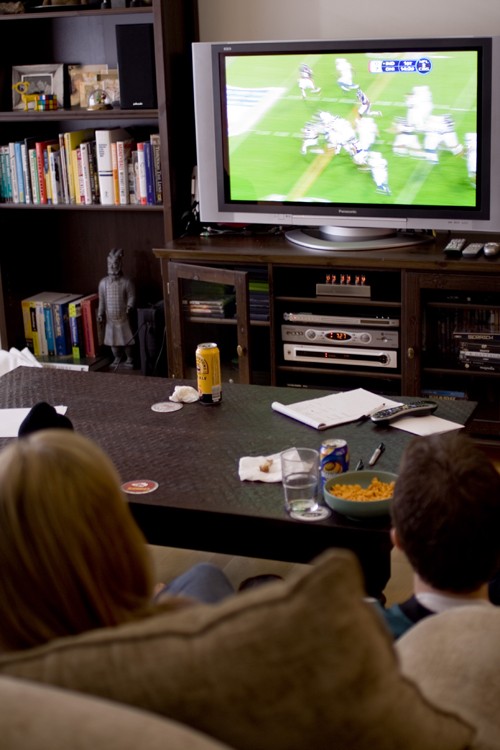 A boy and girl are shown from behind watching a football game on television.