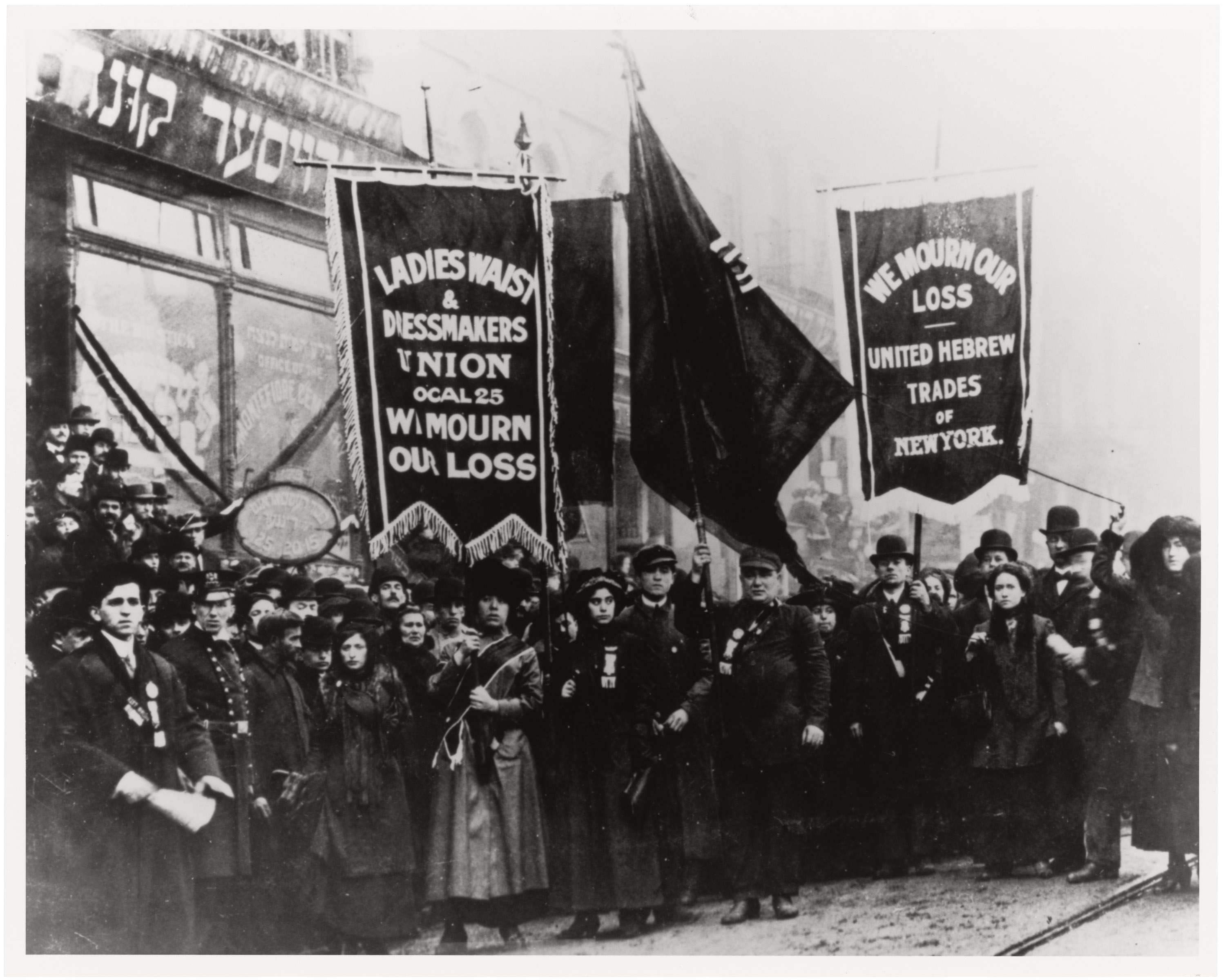 A black and white photo of a crowd of people standing together with large cloth banners. One banner says" Ladies Waist and Dressmakers Union Local 25, We mourn our loss" and the other reads "We mourn our loss, United Hebrew trades of New York".