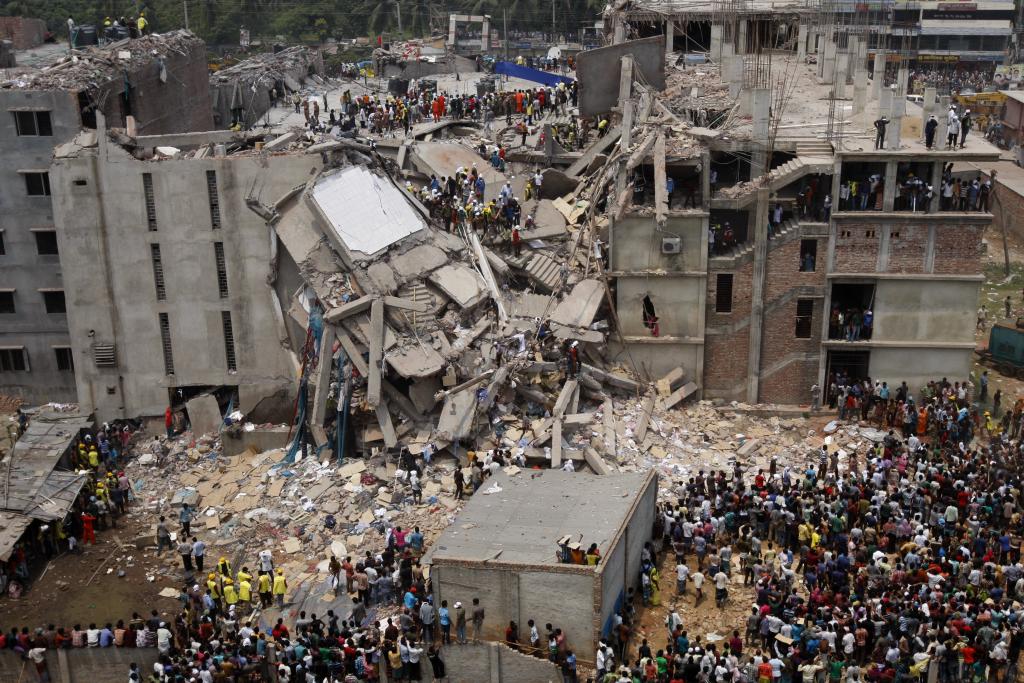 Photograph of collapsed concrete, 5-story building, with people gathered around. The center of the building is entirely caved in with rubble.