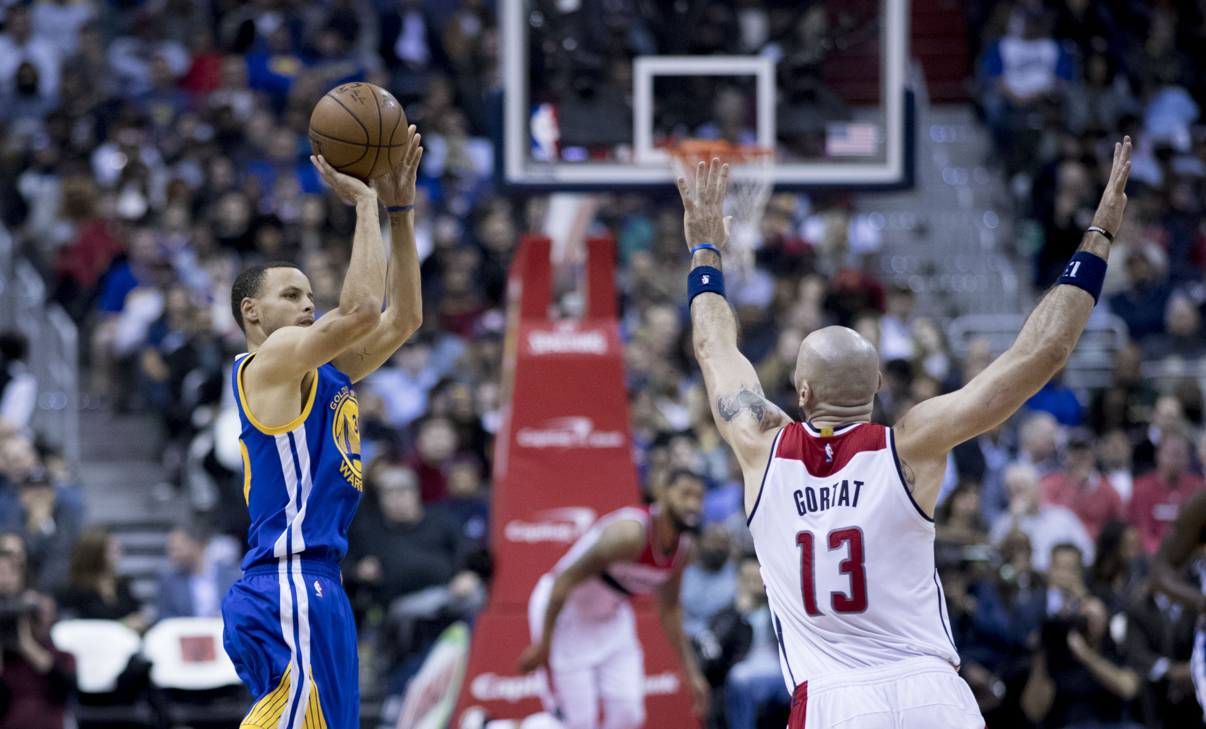In the foreground there are two basketball players. Stephen Curry is on the left and jumping into the air with the basketball in his hands. Marcin Gortat is on the right, facing away from the camera and his arms are in the air as he runs towards Stephen.