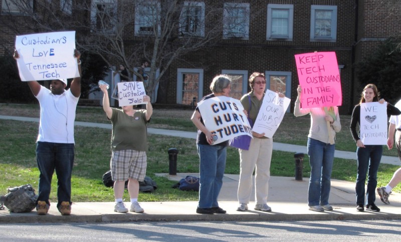 A group of people are shown standing on a sidewalk holding protest signs. Some signs read, "Cuts hurt students" and "Keep tech custodians in the family"