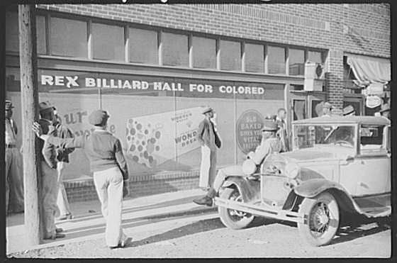 A group of black men and an old car standing outside a billiard hall. The words "Rex Billiard Hall For Colored" are seen on the windows of the hall.