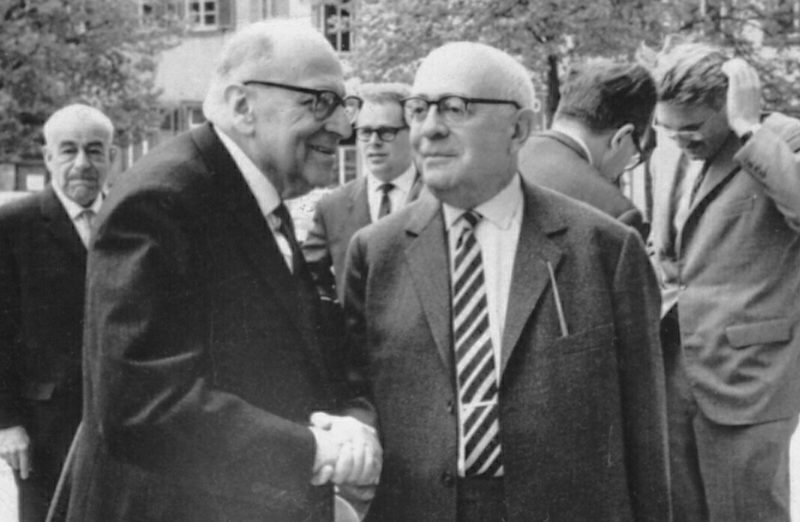 Two elderly men are standing in the foreground of this black and white photo shaking hands.